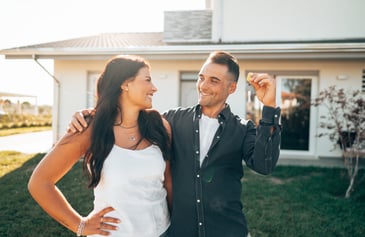A couple purchasing their first home is pictured with keys while standing in their new front yard.