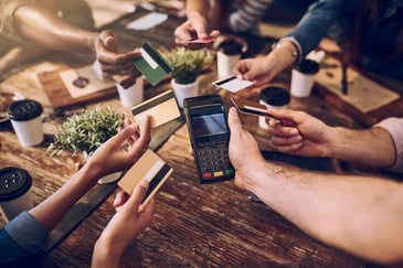Many hands at a restaurant table hold out credit cards to a POS terminal