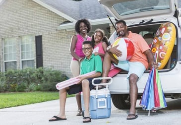 A family sits on the trunk of their car, holding beach supplies.