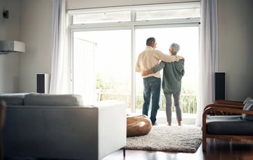 A mature couple stands arm in arm while looking out the window