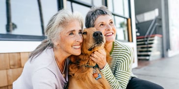 Two women looking on fondly and cuddling with their dog