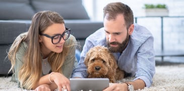 A couple lies on their floor looking at their tablet, with the man holding their dog
