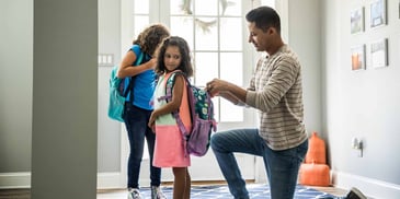 A father helps his young daughter pack her backpack for school