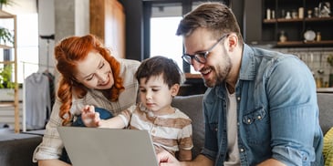 A mother and father sit on their sofa looking at a laptop with their young son