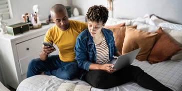A young couple sits on their bed, looking at financial notes in a book and on their phone and laptop