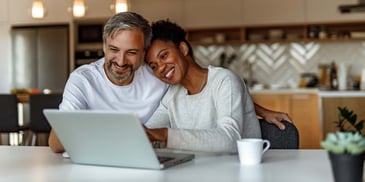 A couple smiles and holds each other while looking at a laptop in their kitchen