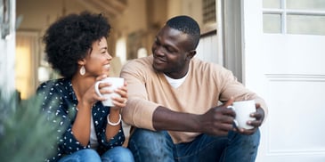 A couple sits on their patio, smiling at each other and drinking coffee