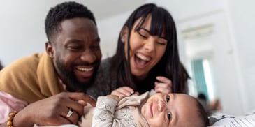 A young couple smiles and looks on their infant child, who is also smiling