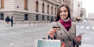 A smiling woman out on the town square, looking at her credit card while holding shopping bags