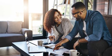 A couple sits in their living room smiling at each other, as they work on finances