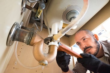 A plumber works on pipes under a sink