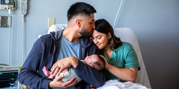 A father kisses the head of his partner as he holds their newborn baby in the hospital delivery room