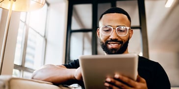 A smiling young man with glasses sits on a couch working on a tablet