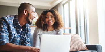 A smiling couple sit in their living room, using a laptop to go over finances