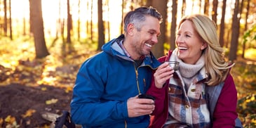 A couple sits on a bench in the woods during the fall season, smiling at each other