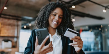 A woman smiles while holding her phone in one hand and credit card in the other