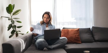 A young woman sits on her sofa with her laptop, reviewing a paper document