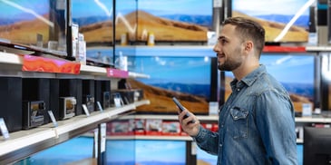 A young man holding his cell phone and looking at televisions for sale in a store