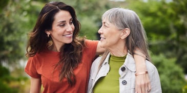 An adult daughter places her arm around her mother, the two smiling at each other