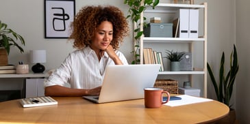 A woman sitting at a table using her laptop, resting her head on her hand