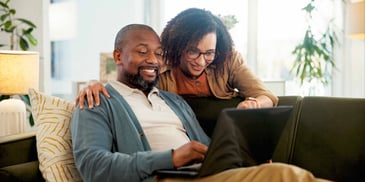 A woman with her arm around a man and pointing at something on the laptop in his lap