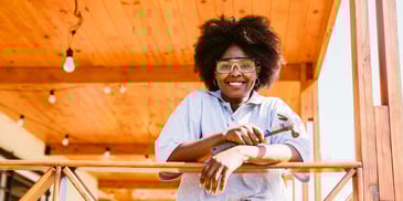 A woman stands on the deck outside her home, wearing safety goggles and holding a hammer
