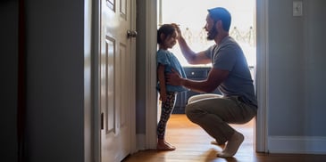 A father crouches down, measuring his young daughter's height against the door frame