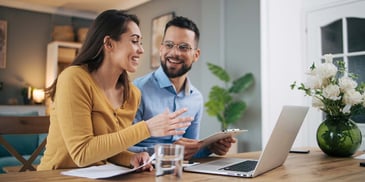 A young couple sit at their dining room table with a laptop in front of them, reviewing finances