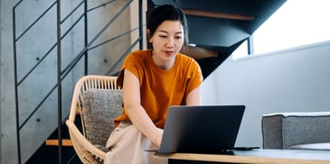 A woman sits in a chair and leans forward, typing on her laptop placed on a table in front of her