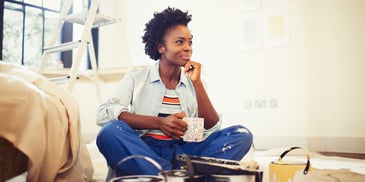 A woman sitting on the floor of her home amongst paint supplies, looking thoughtful