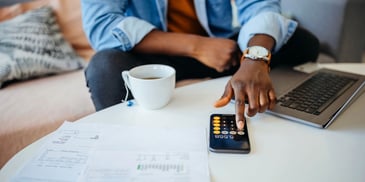 A hand works on the calculator app of a mobile phone, with an open laptop and coffee cup nearby