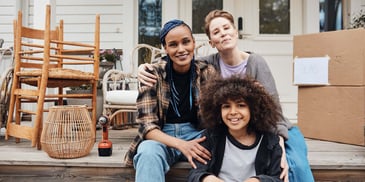 A smiling couple sits on their porch with their son, surrounded by moving boxes