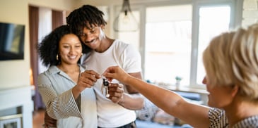 A young couple smiles and stands closely together, as a woman with an outstretched hands gives them keys to their new home