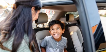 A young boy smiles up at his mother, as he's put into a booster seat in their car