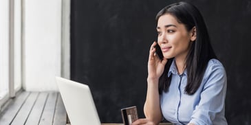 A woman sits at a desk with her laptop on a phone call, holding out her credit card in one hand