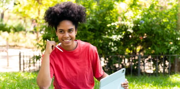 A young woman sitting in a park, smiling and using a tablet to do finances