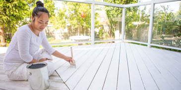 A woman sits on her outdoor deck, painting the wood white