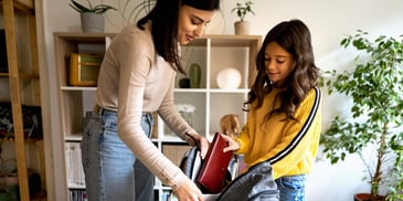 A mother helps her daughter pack her backpack as she prepares to head to school