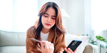 A young woman in a sweater sits on a couch, her phone in one hand and her credit card in the other hand