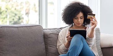A woman sits on the sofa in her living room, looking down at her tablet while holding out a credit card