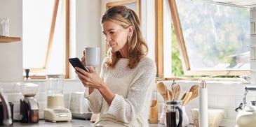 A woman stands in her kitchen against the sink, holding a coffee mug in one hand and a cell phone in the other