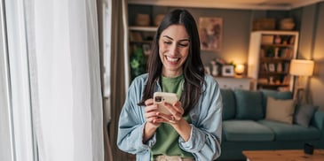 A young woman stands in her living room and smiles while looking at her cell phone