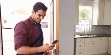 A young man smiles and walks into his home, holding his mobile phone in his hand