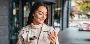 A young woman walking outside a storefront smiles looking down at her mobile phone and holding a coffee