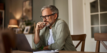 A man sits at his dining room table and looks thoughtfully at his laptop screen