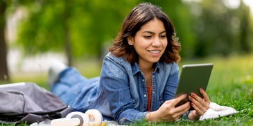 A young woman lays down in a park, smiling and using a tablet with a pair of headphones beside her