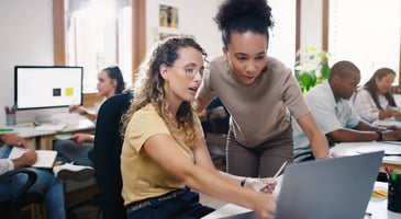 A manager stands next to an employee at her desk to provide guidance