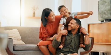A young, smiling family sits together in their living room