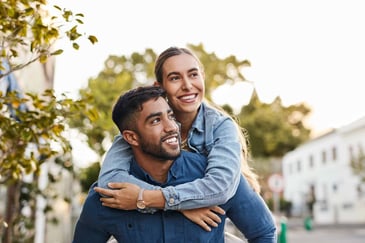 A young woman piggyback rides on her partner, both smiling.
