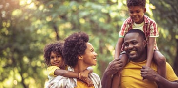 A family of four smiles at one another as the parents carry the children in a park.